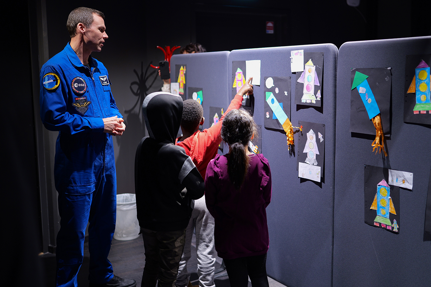 Tre barn visar teckningar för en man i astronautdräkt. 