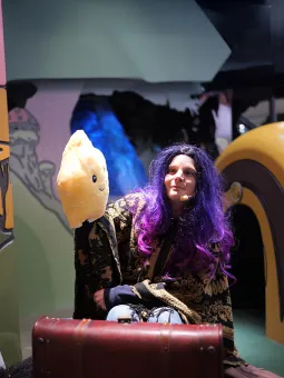 En kvinna med peruk sitter framför en resväska och håller upp en gosedjursstjärna.