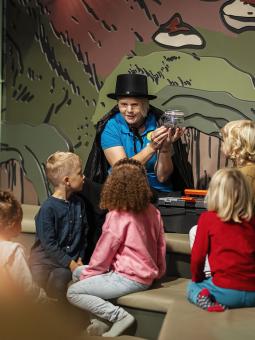 En kvinna i hatt och cape visar upp en glasburk med ett läskigt föremål för en grupp av barn.
