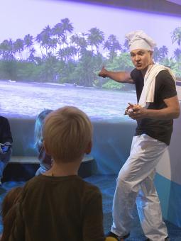 En man utklädd med kockmössa håller i en föreställning för en publik med barn och vuxna.