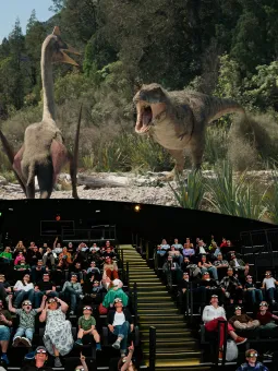 En blandad publik sitter i mwisdome och stirrar upp mot en dinosaurie som syns på duken.