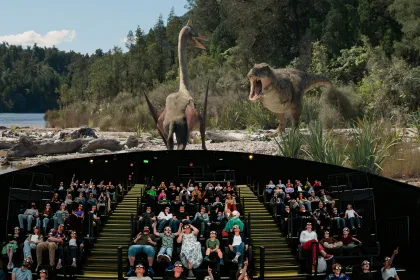 En blandad publik sitter i mwisdome och stirrar upp mot en dinosaurie som syns på duken.